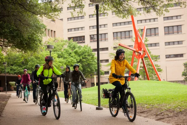 A group of cyclists bike along campus, away from a large red metal sculpture. 
