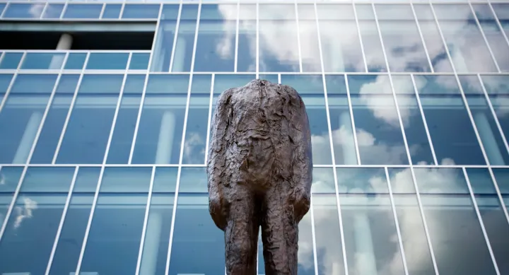 A headless bronze statue behind a building made of panes of glass which reflect the sky.
