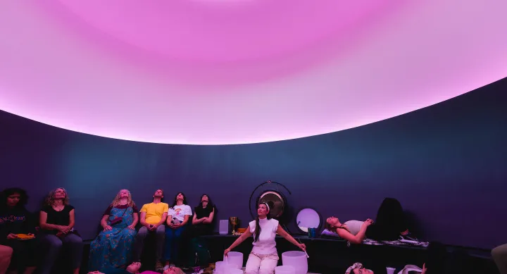 A woman sits in a curved room with purple lighting and an oculus opening to a white sky above her. While she singing bowls of various sizes, she is surrounded by a group of about 15 people who are all looking up.