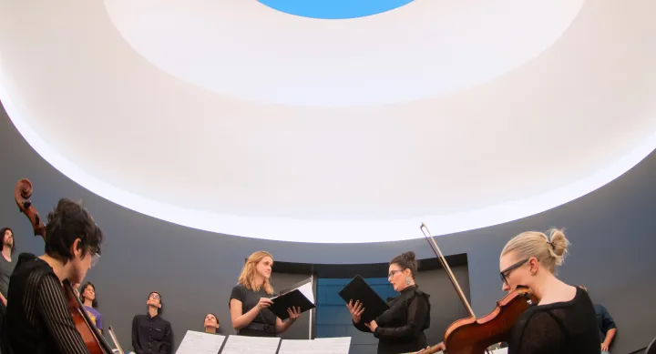 Two singers perform in a curved room while two string musicians preform next to them. All are in a curved room with an oculus in the roof.