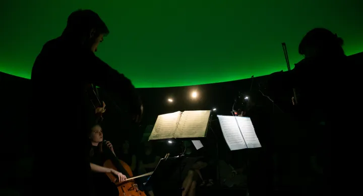 The silhouette of two violinists performing in a dark curved room lit by a green light.