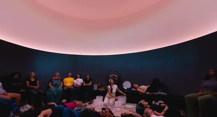 A women plays singing bowls while surrounded by sitting and laying listeners. She sits in a curved room lit with pink light underneath an oculus in the ceiling.