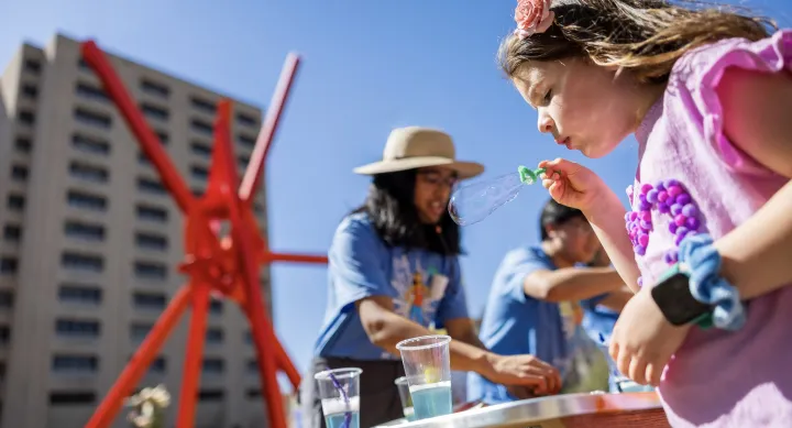 A young girl blows a soap bubble outside in front of a large red metal sculpture.