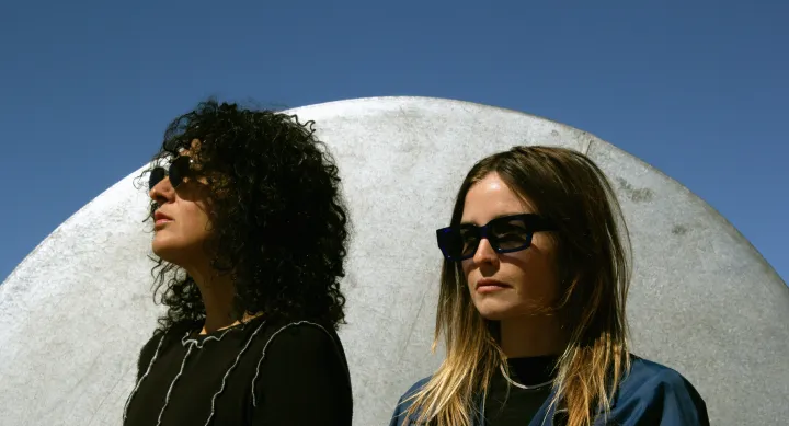A portrait of two women standing side by side wearing sunglasses. They are outside, and behind them is a cement dome.
