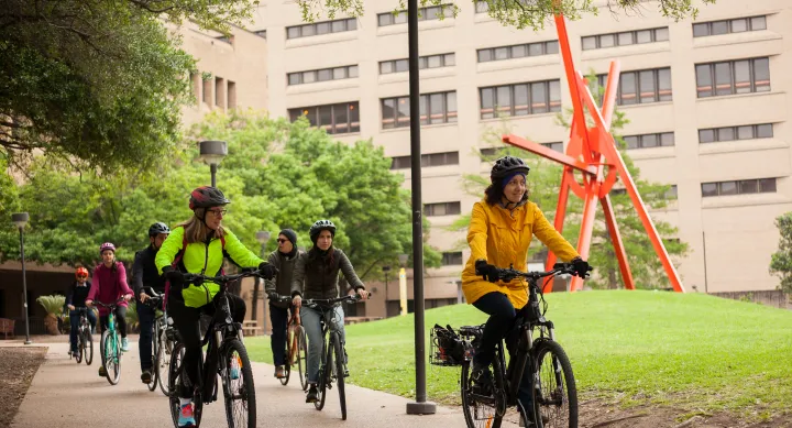 A group of cyclists bike along campus, away from a large red metal sculpture. 