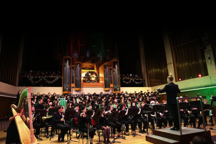 The University of Texas Wind Ensemble plays with combined choirs, conducted by J.D. Burnett. Bates Recital Hall is decorated with lights and Christmas garland.