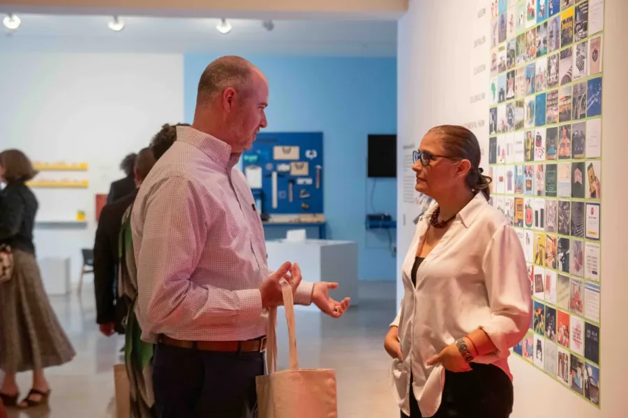 Two attendees talk during an MFA info session, standing in front of a wall filled with colorful graphic design tiles. The interaction takes place in a well-lit gallery space with other guests exploring the exhibition in the background.