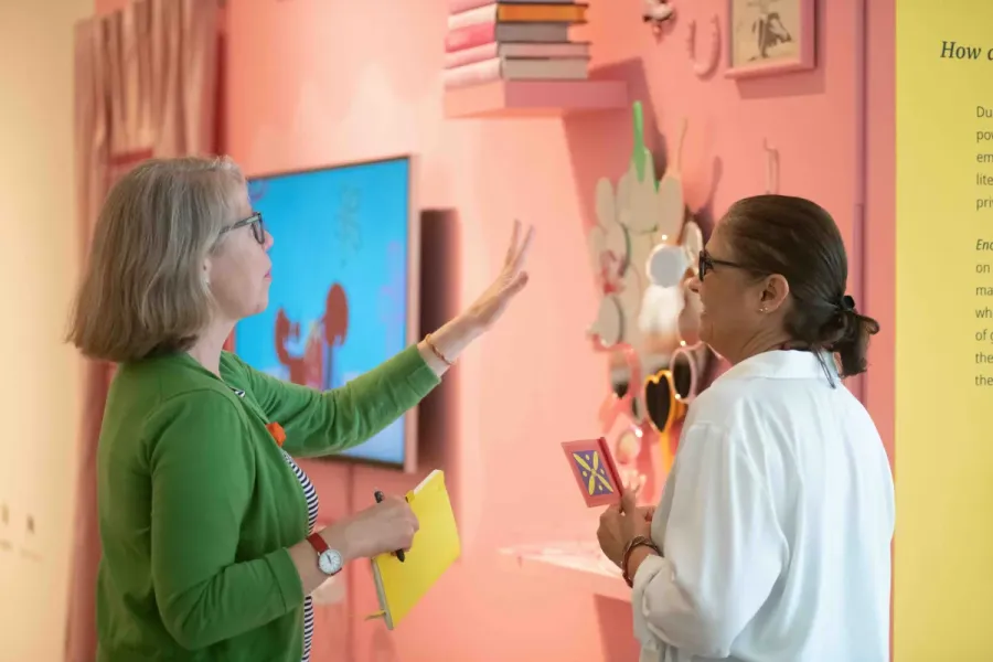 Two attendees chat in front of a pink wall display featuring colorful mirrors and floating books. One gestures with her hand while holding a notebook, and the other smiles while holding a brightly designed card.