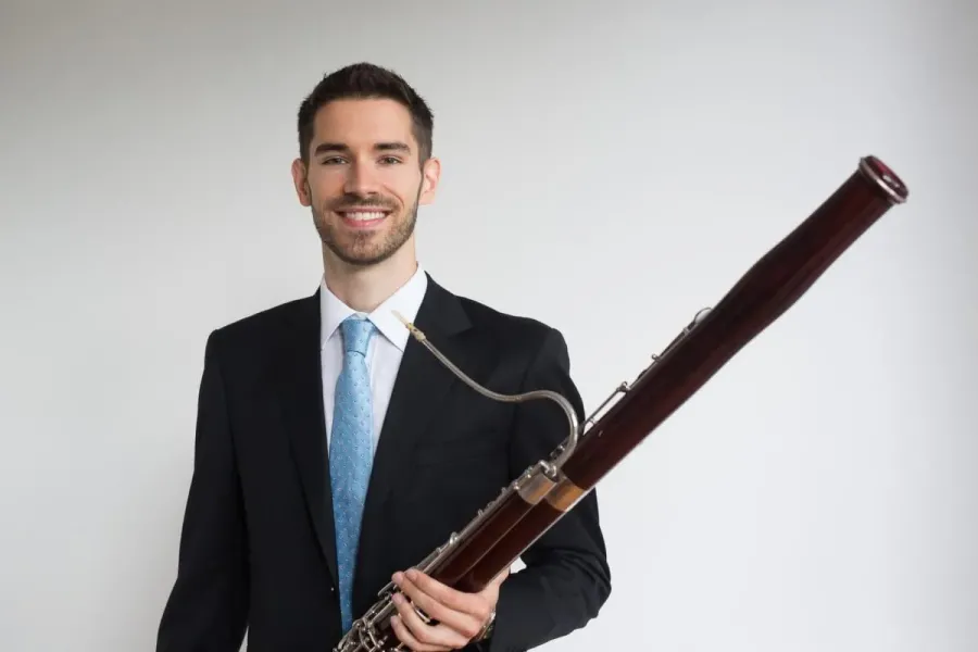 Drew Pattison holds his bassoon wearing a black suit with a blue tie.