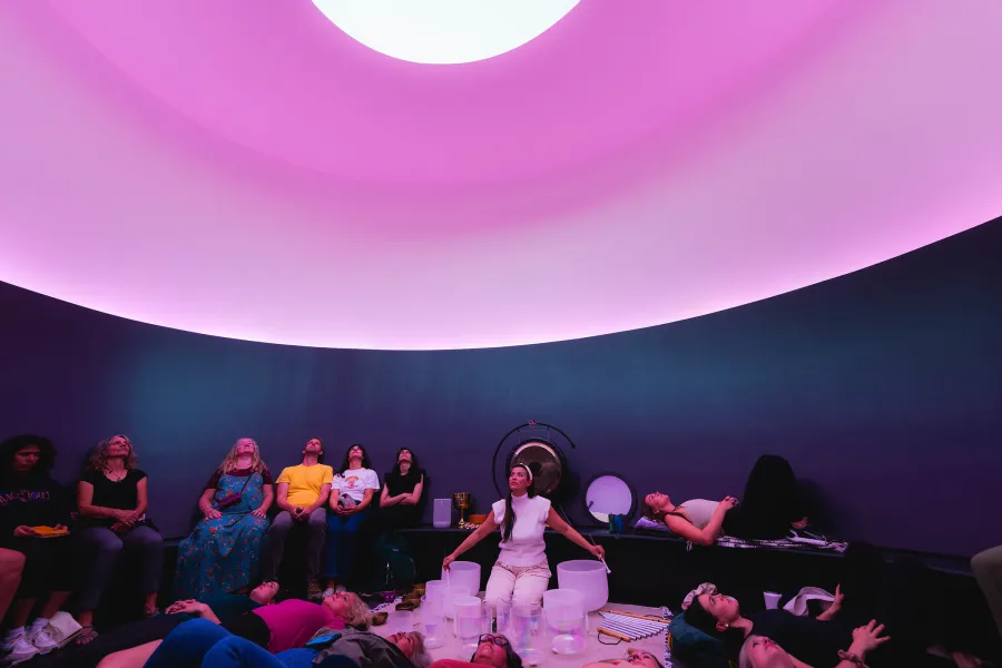 A woman sits in a curved room with purple lighting and an oculus opening to a white sky above her. While she singing bowls of various sizes, she is surrounded by a group of about 15 people who are all looking up.