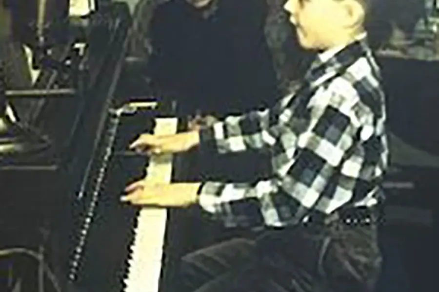 A very young Gregory Allen plays an upright piano while a woman looks on. 