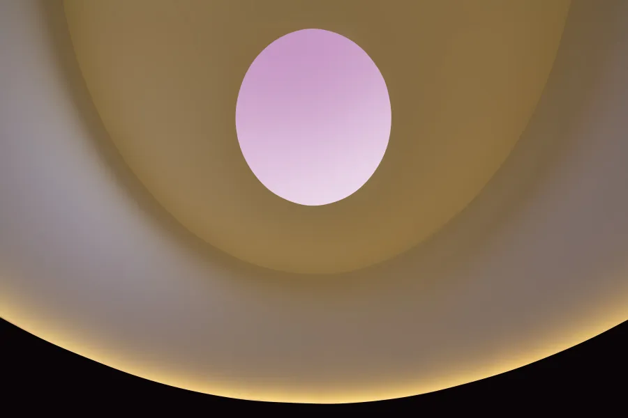 Ceiling of james turrells skyspace, featuring an orange lit wall and an occulus with a pink sky